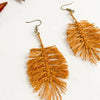 A pair of handmade dangle earrings with textured, fringed leaf-shaped drops in a warm mustard-gold color, attached to silver fishhook wires, displayed on a clean white background.