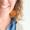 A close-up lifestyle shot of a smiling woman wearing a large, textured mustard-yellow fringe earring. She has curly light brown hair and is wearing a blue denim shirt under a light gray cardigan, set against a clean white background.