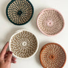 Four mini woven bowls in natural flax and dark green, pale pink, ivory, and rust orange with a hand holding one against a white background.
