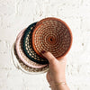 Hand holding a stack of four different colors of woven mini baskets against a light background.