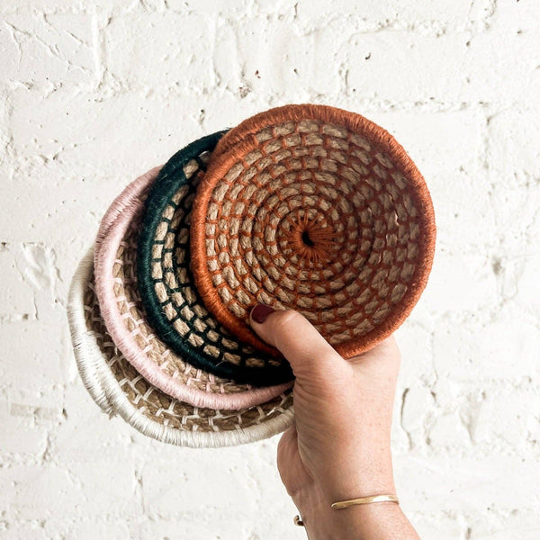 Hand holding a stack of four different colors of woven mini baskets against a light background.