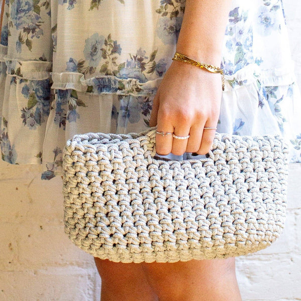 A close-up of a person holding a chunky crocheted clutch bag in a light beige color. The person is wearing a cream-colored floral dress and gold jewelry, standing against a white brick wall.
