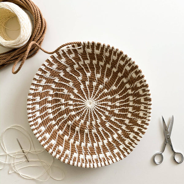 Partially woven basket with brown and white pattern on a light surface, accompanied by scissors, twine, and a darning needle.