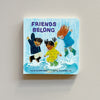 A colorful board book called 'Friends Belong' featuring an illustration of three children in rain gear splashing in puddles. The book rests against a neutral backdrop.