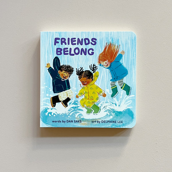 A colorful board book called 'Friends Belong' featuring an illustration of three children in rain gear splashing in puddles. The book rests against a neutral backdrop.