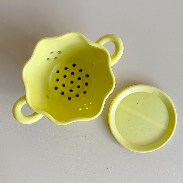 Top-down view of a yellow ceramic berry bowl colander and matching drip plate on a neutral background