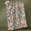A tea towel in white with orange, green, and brown botanical designs hanging from a wire against a green wall.