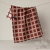 The reverse side of a kitchen towel showing the contrasting dark red with cream grid plaid pattern, hanging from a wire next to a glass carafe and cup.