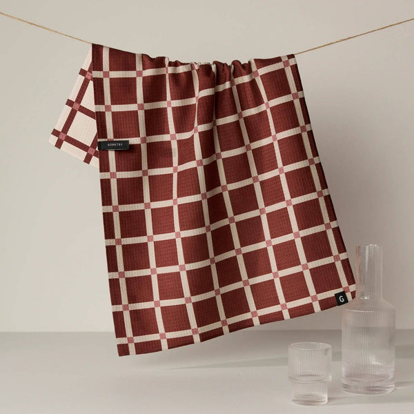 The reverse side of a kitchen towel showing the contrasting dark red with cream grid plaid pattern, hanging from a wire next to a glass carafe and cup.