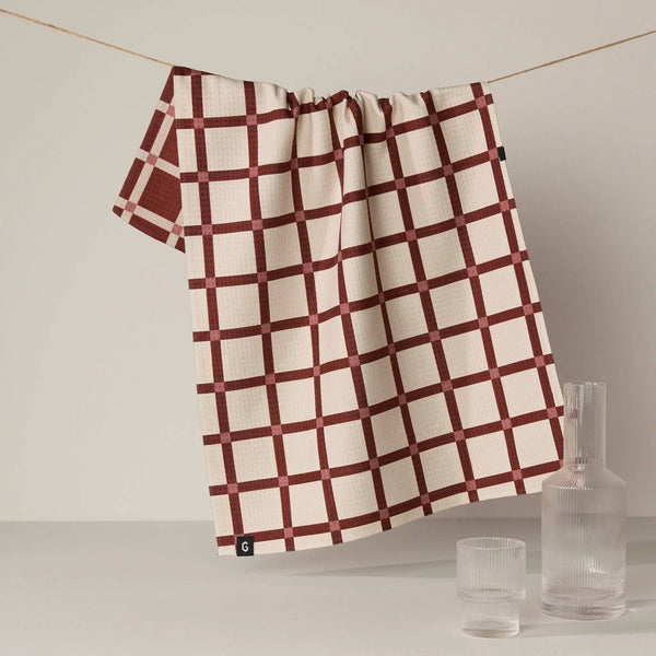 A kitchen towel in cream and dark red grid plaid hangs from a wire against a soft neutral backdrop next to a clear glass carafe and cup with water.