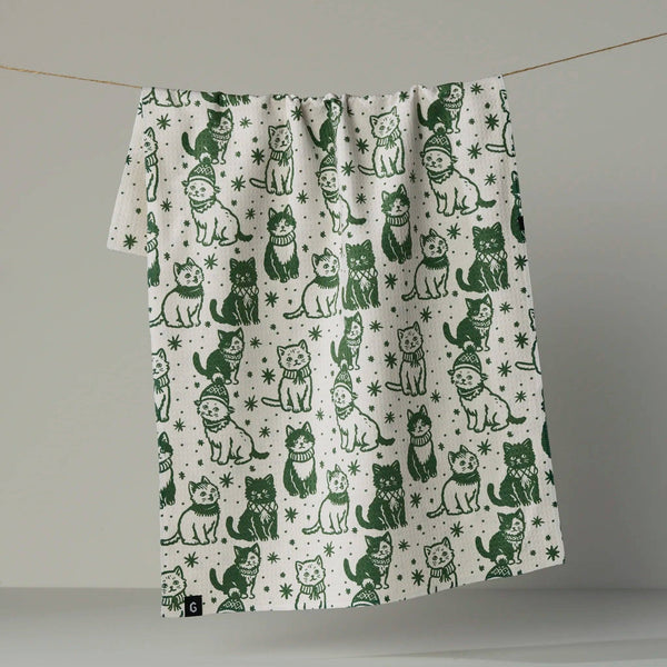 A white kitchen towel printed with a green pattern of cats, hanging from a thin string against a neutral gray background and revealing the reverse side, which has a print of tiny stars.