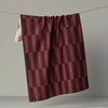 A dark red tonal geometric patterned tea towel hanging from a wire against a neutral backdrop, partially revealing the light botanical patterned reverse side. 