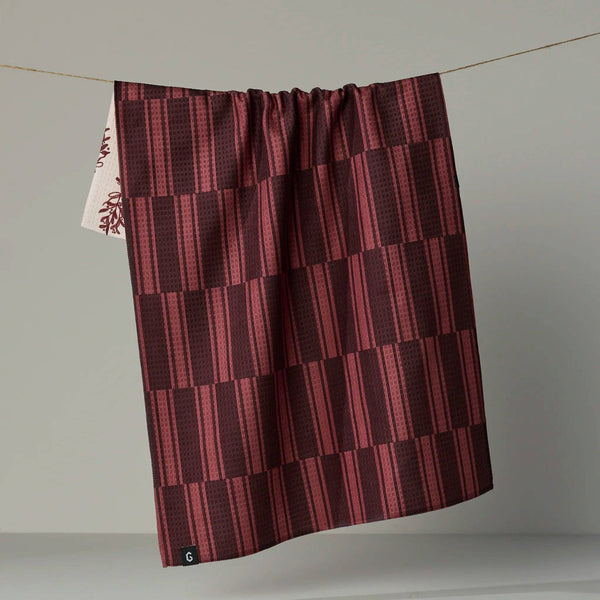 A dark red tonal geometric patterned tea towel hanging from a wire against a neutral backdrop, partially revealing the light botanical patterned reverse side. 