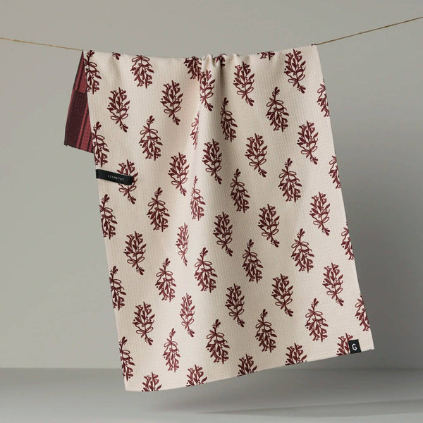 A cream-colored kitchen towel with a repeating dark red botanical block print pattern, hanging from a thin wire on a neutral gray background, partially revealing the dark red geometric pattern on the reverse side.