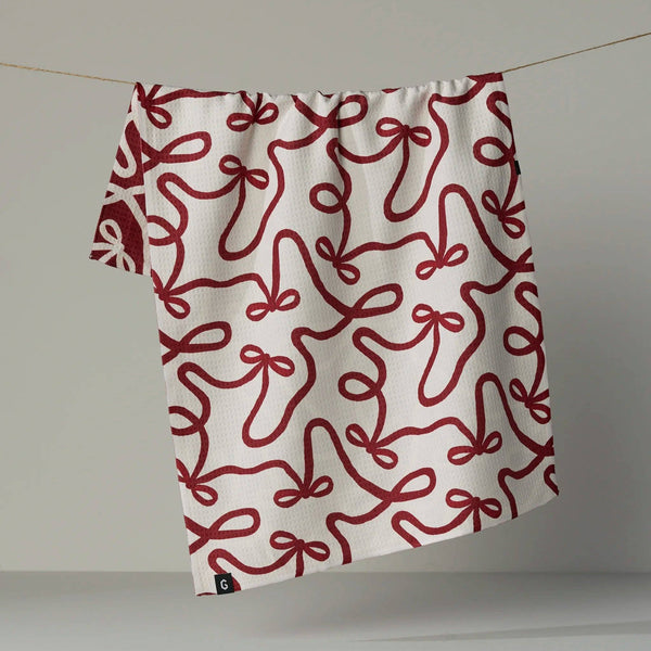 A white kitchen towel with a red bow pattern hanging from a white, revealing the reverse side in the same print in a reverse colorway, photographed against a neutral backdrop.