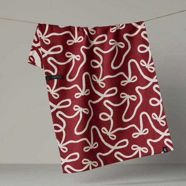 A red kitchen towel with a white bow pattern hanging from a white, revealing the reverse side in the same print in a reverse colorway, photographed against a neutral backdrop.