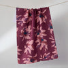 A tea towel hanging from a thin cord against a plain light gray wall. The towel has a waffle texture and features a floral pattern in dusty pink, mauve, and dark navy blue.