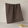 A dark brown kitchen towel with a white cross pattern hangs from a string, with the reverse white patterned side partially visible behind it. A wooden scrubber rests on a small dish in the foreground against a neutral background.