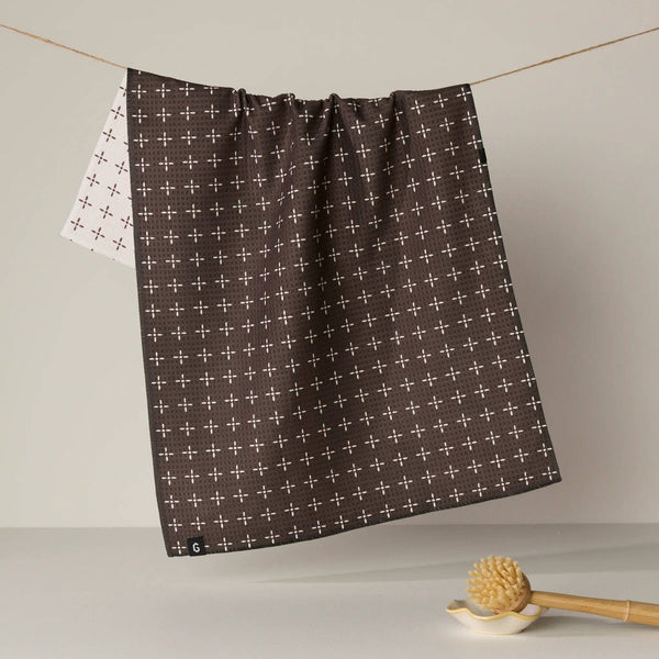 A dark brown kitchen towel with a white cross pattern hangs from a string, with the reverse white patterned side partially visible behind it. A wooden scrubber rests on a small dish in the foreground against a neutral background.