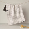 A white kitchen towel with a brown cross pattern hangs from a string on a line, with the back side showing a brown cross on white pattern. A wooden scrubber rests on a small white dish in the foreground against a neutral background.