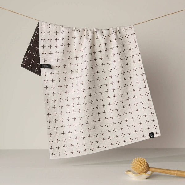 A white kitchen towel with a brown cross pattern hangs from a string on a line, with the back side showing a brown cross on white pattern. A wooden scrubber rests on a small white dish in the foreground against a neutral background.