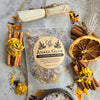 A clear stand-up pouch labeled 'Gnat and Bee Amber Glow Botanical Bath Soak' tied with twine at the top, surrounded by dried orange slices, cinnamon sticks, loose yellow flowers, and a small bowl on a light textured surface.
