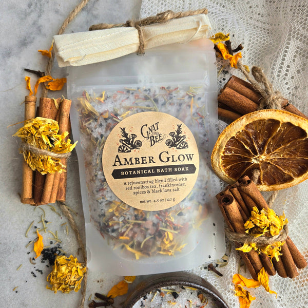 A clear stand-up pouch labeled 'Gnat and Bee Amber Glow Botanical Bath Soak' tied with twine at the top, surrounded by dried orange slices, cinnamon sticks, loose yellow flowers, and a small bowl on a light textured surface.