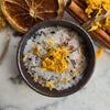 A dark ceramic bowl filled with coarse salt crystals and dried yellow flowers, styled on a light marble surface with a dried orange slice, cinnamon sticks, and loose flower petals around it.