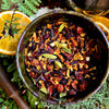 A close-up overhead shot of a dark ceramic bowl filled with loose-leaf mulling spice blend. The blend contains dried flowers, citrus peel, green cardamom pods, and whole spices.