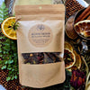 A kraft paper stand-up pouch of Gnat and Bee Blood Moon Mulling Spices, surrounded by dried orange slices, green herbs, cinnamon, and a metal mug on a rustic styled background.