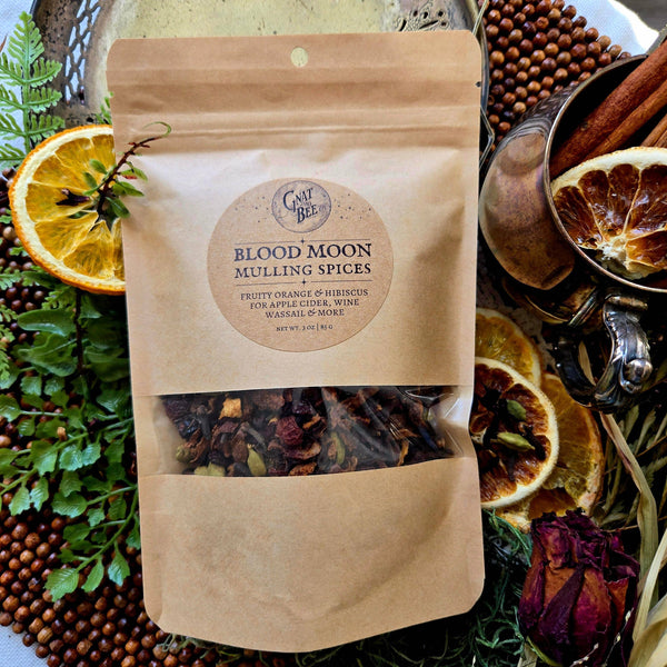 A kraft paper stand-up pouch of Gnat and Bee Blood Moon Mulling Spices, surrounded by dried orange slices, green herbs, cinnamon, and a metal mug on a rustic styled background.