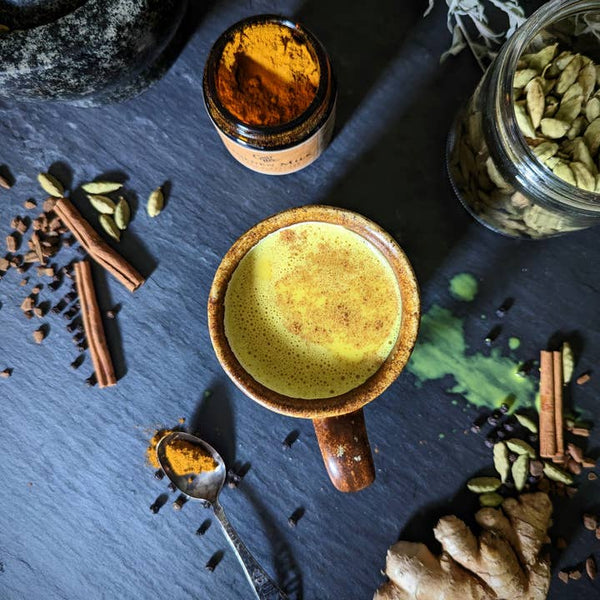 A brown ceramic mug filled with golden milk on a grey background. It is surrounded by a jar of golden milk blend and loose spices.