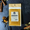 A sample-sized kraft packet of golden milk blend on a grey background surrounded by spices and a silver spoon. 