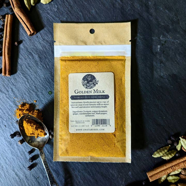 A sample-sized kraft packet of golden milk blend on a grey background surrounded by spices and a silver spoon. 