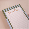 A close-up of a to-do list notepad with a striped border in sage green and light blue, featuring the words 'to do list' in brown script on a lined white page, set against a textured beige background.