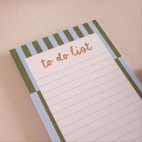 A close-up of a to-do list notepad with a striped border in sage green and light blue, featuring the words 'to do list' in brown script on a lined white page, set against a textured beige background.