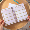Hands holding a spiral-bound planner with a reading progress tracker on a wooden surface