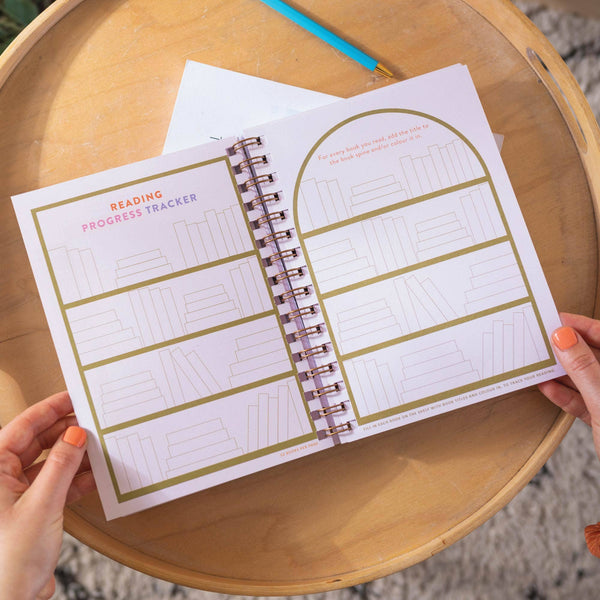 Hands holding a spiral-bound planner with a reading progress tracker on a wooden surface