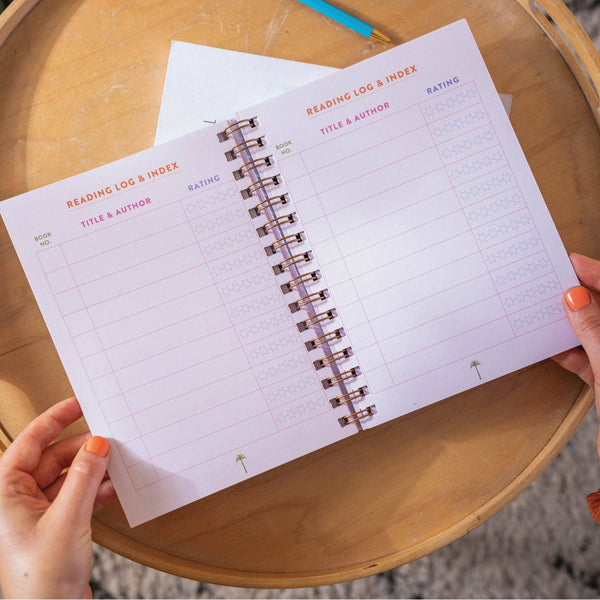 Hand holding a spiral-bound notebook with a reading log and index on a wooden surface