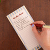 A close-up of a hand writing on a lined notepad labeled 'to do list' with a red pen, placed on a dark wood surface.