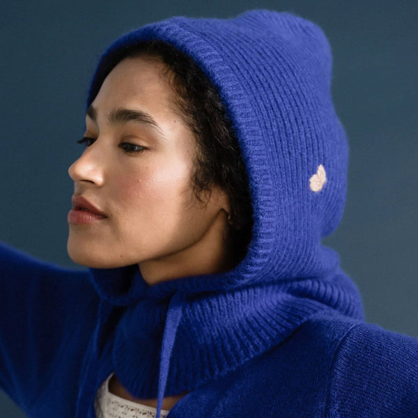 A person with dark hair wearing a blue sweater over a white top along with a blue knit balaclava hoodie, accented with a beige embroidered heart, standing against a shadowy blue backdrop.