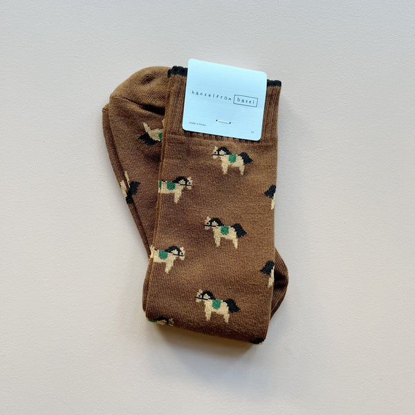 A pair of tall crew socks with 'hansel from basel' packaging featuring a brown earth toned design of equestrian horses against a neutral background. 