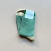 A pair of crew socks with 'hansel from basel' packaging in a marled green and beige ribbed knit with a solid green top stripe and beige heels and toes laying against a neutral background.