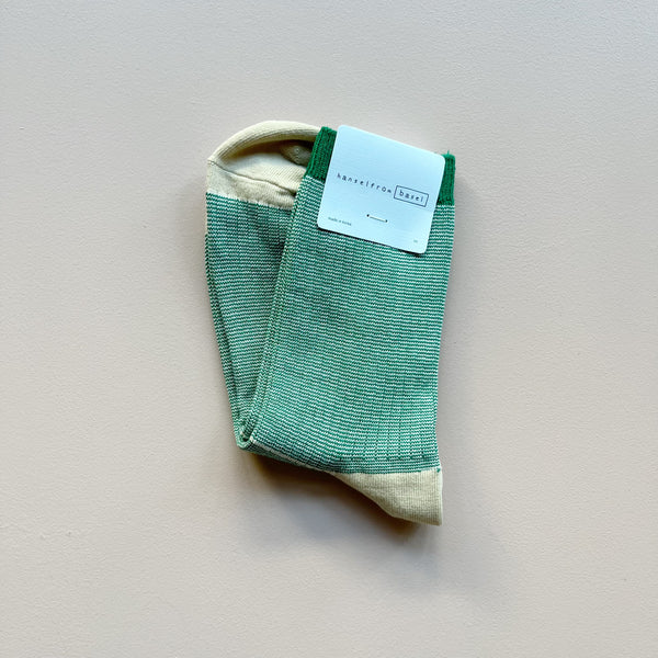 A pair of crew socks with 'hansel from basel' packaging in a marled green and beige ribbed knit with a solid green top stripe and beige heels and toes laying against a neutral background.