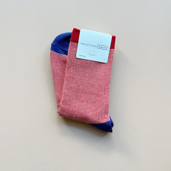 A pair of crew socks with 'hansel from basel' packaging in a marled red and white ribbed knit with a solid red top stripe and cobalt blue heels and toes laying against a neutral background.