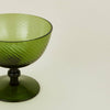 A close-up view of an olive green coupe glass with a textured, ribbed exterior and a dark green stem and base, photographed on a clean, solid off-white background.