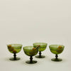 Four green glass coupe-style cocktail glasses with stems, arranged on a plain light background. Each glass contains a cocktail with a few olives on toothpicks.