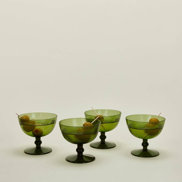Four green glass coupe-style cocktail glasses with stems, arranged on a plain light background. Each glass contains a cocktail with a few olives on toothpicks.