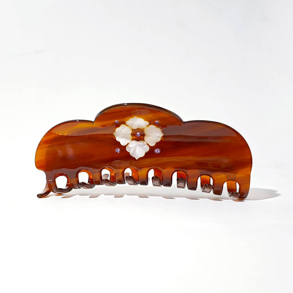 A glossy tortoiseshell hair claw clip in a warm amber-brown tone, decorated with a central white floral motif and small purple rhinestone accents, photographed on a clean white background.