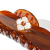 A detailed view of a glossy amber-brown hair claw clip with a scalloped edge, featuring a white floral embellishment and small purple rhinestone accents, shown on a white background.